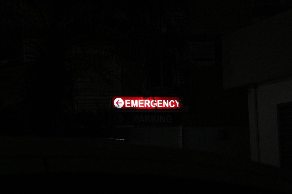 Czerwony, podświetlany znak „Emergency” wskazujący kierunek wyjścia awaryjnego w ciemnej przestrzeni.