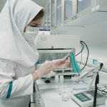 Pracownica laboratorium w maseczce i rękawiczkach analizuje próbkę kosmetyku przy użyciu sprzętu laboratoryjnego.